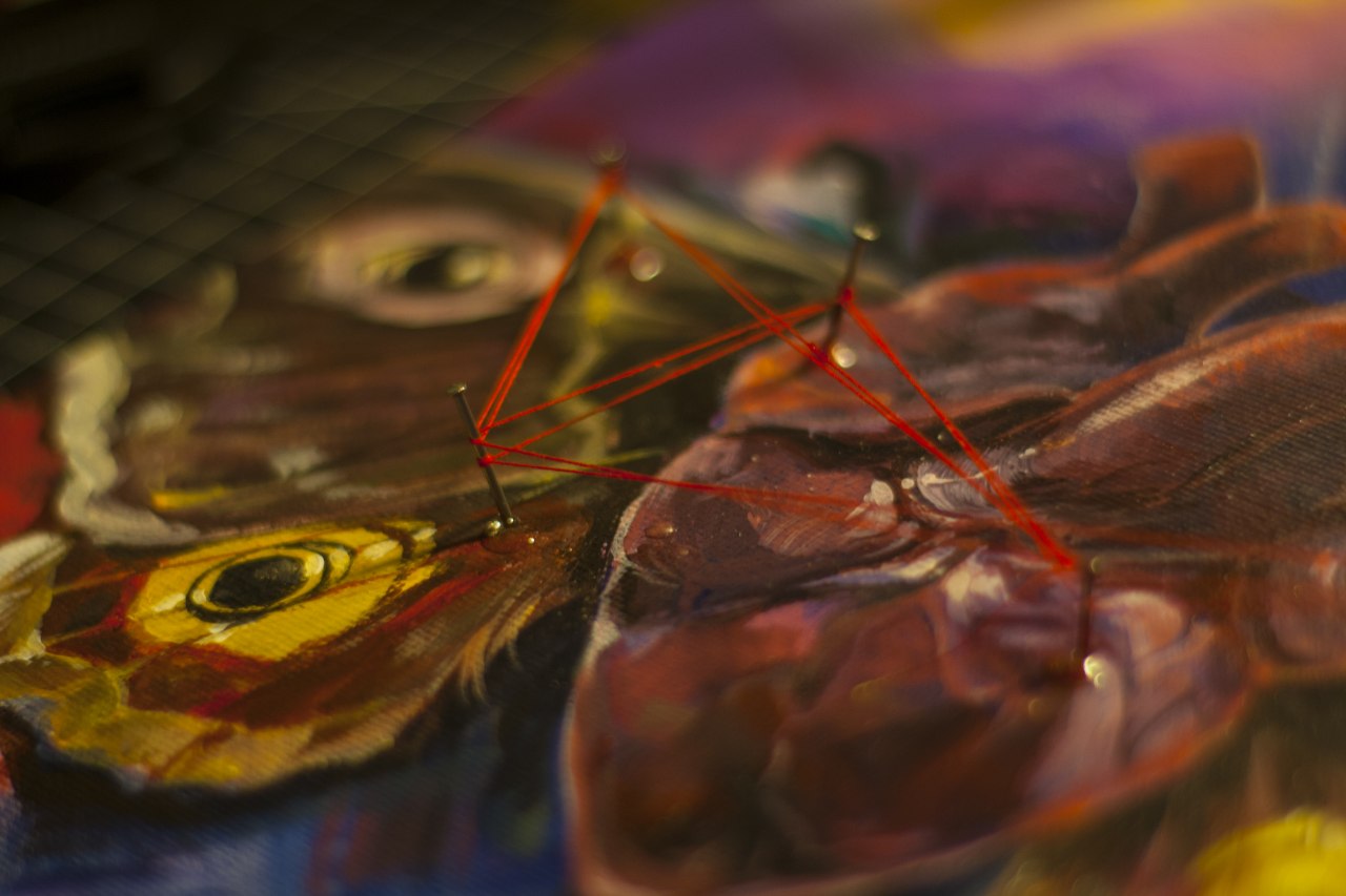 an angled shot of an acrylic painting of a human heart with moth wings, perforated by small nails connected with a red string